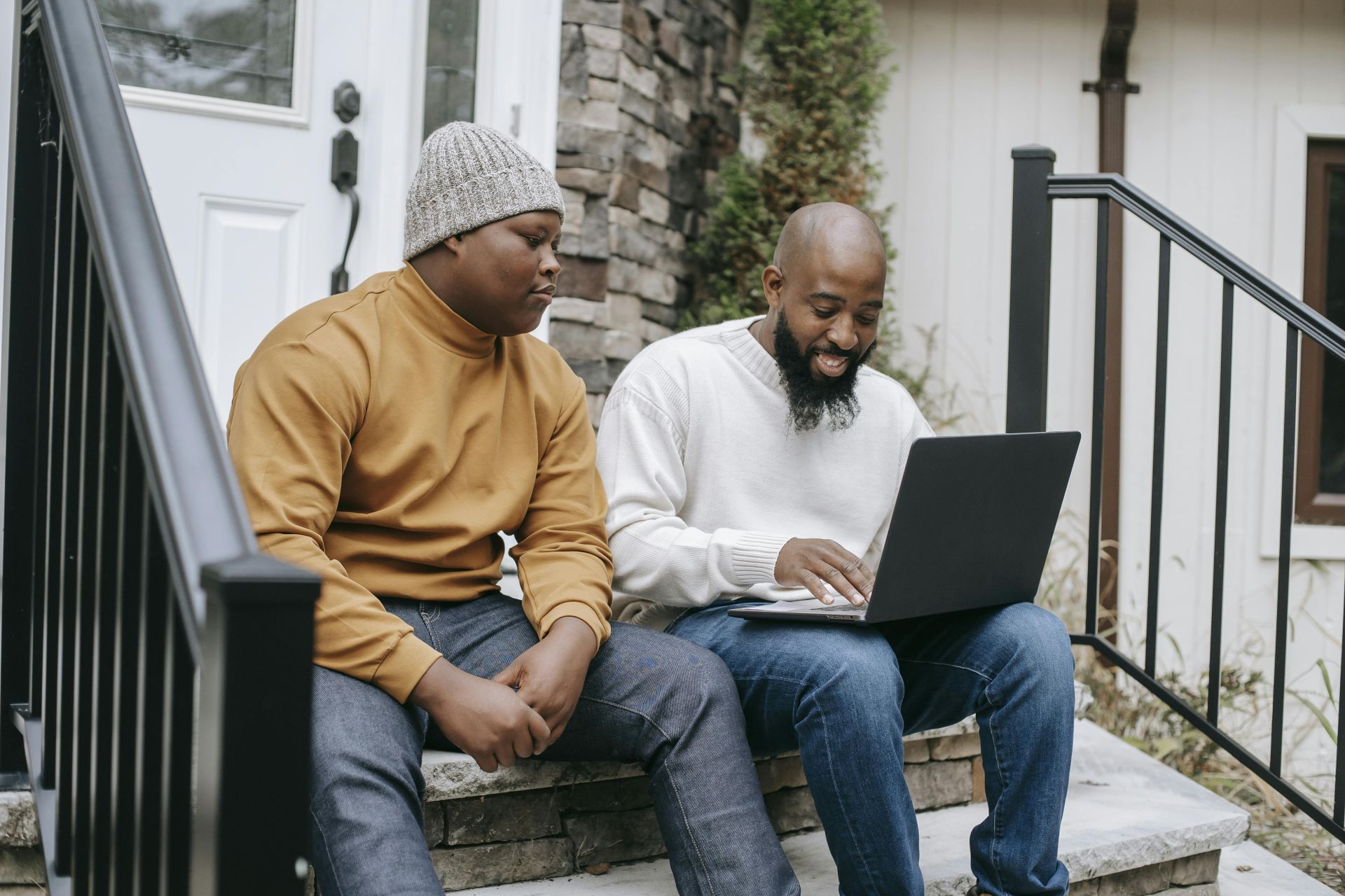 Positive bearded African American male remote worker sitting on stairs and typing on netbook keyboard with teen son