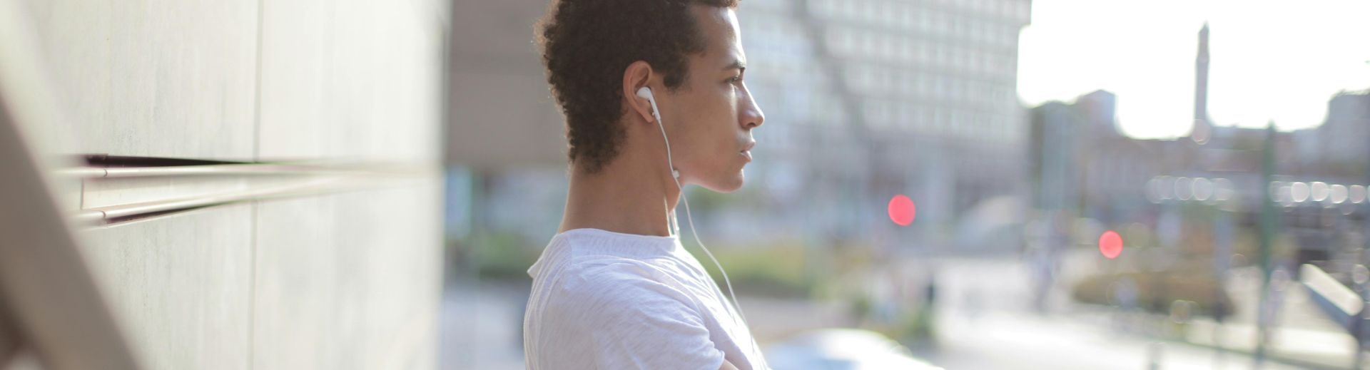 Side view of calm thoughtful African American male athlete in earphones and sportswear looking away and stretching leg while standing alone and listening to music on street near modern building in city